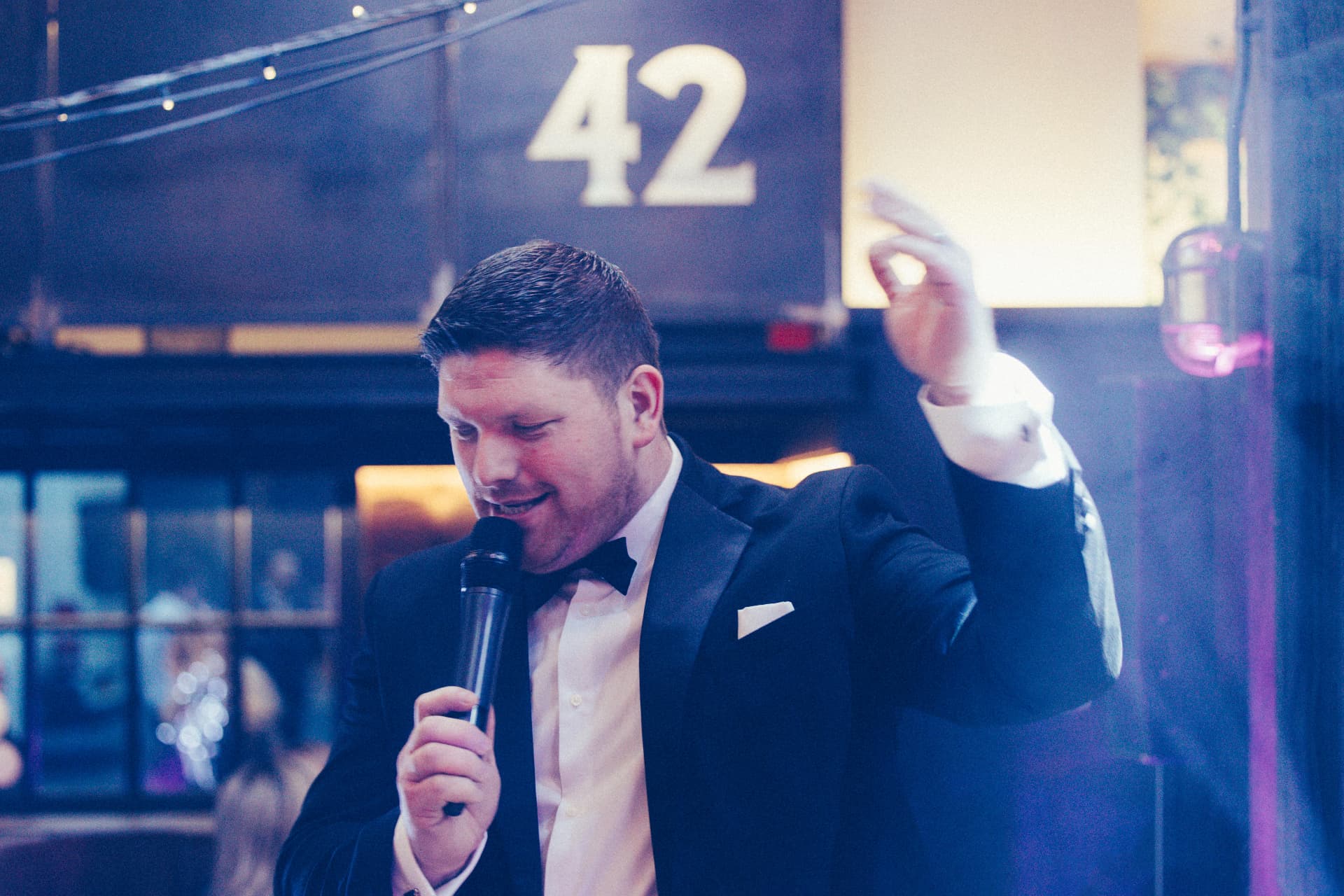 Jon Kane speaking into a microphone at a wedding reception, one hand raised, under coloured party lighting