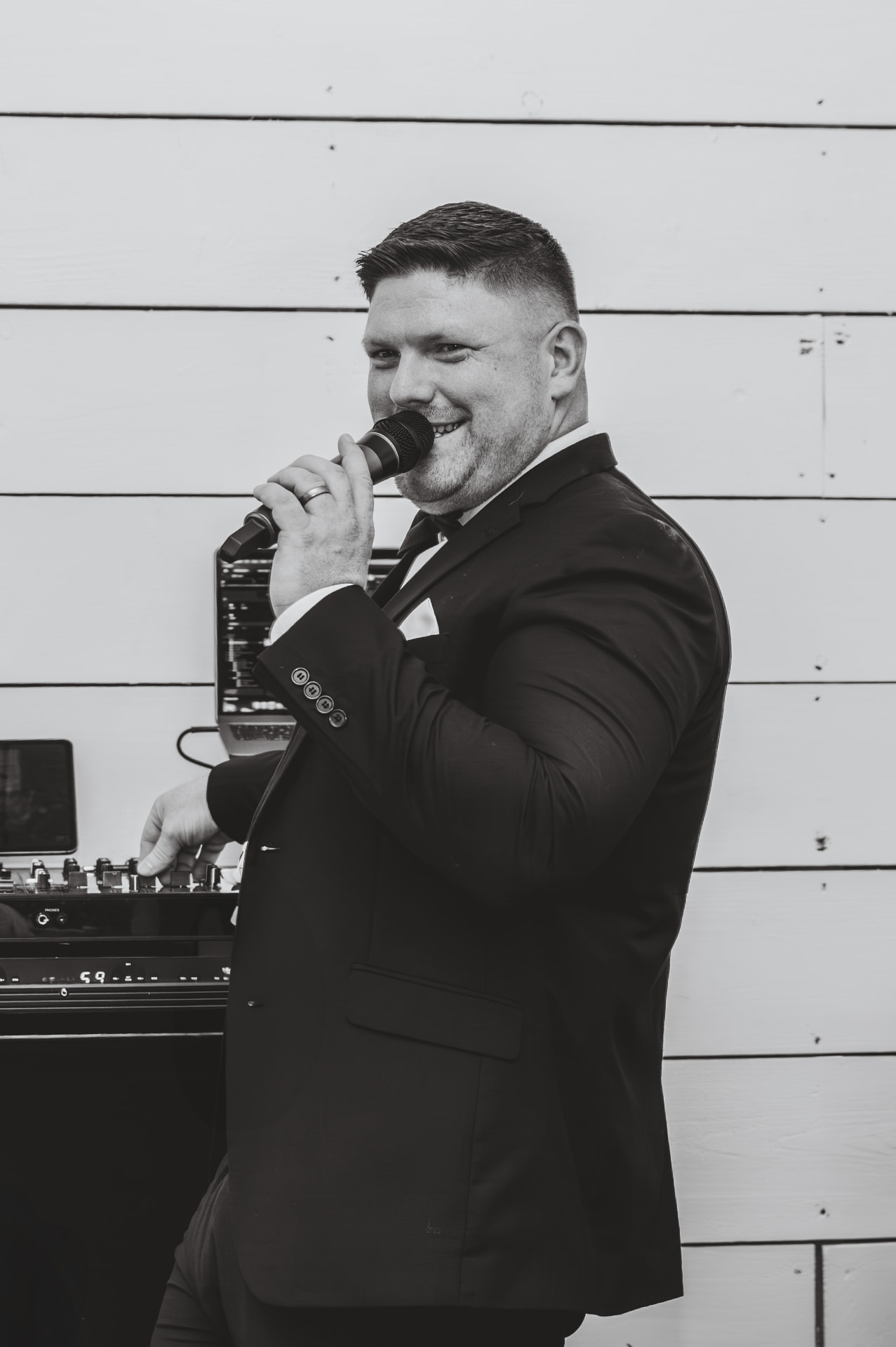 Jon Kane in a tuxedo and bow tie, microphone in hand, at the DJ decks